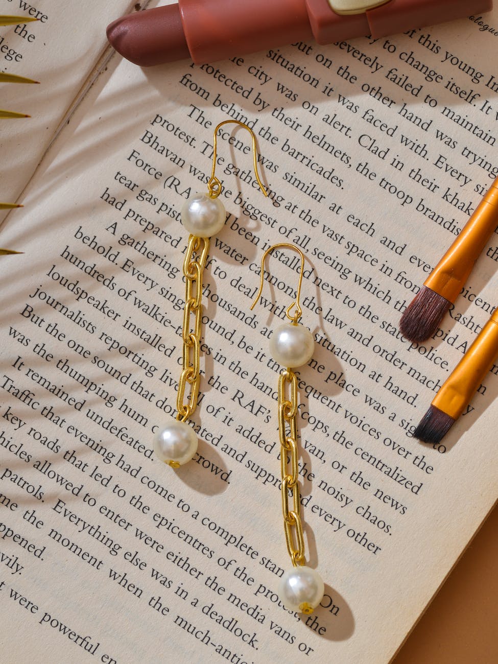 chain gold and pearl earrings on a book beside a lipstick and make up brushes